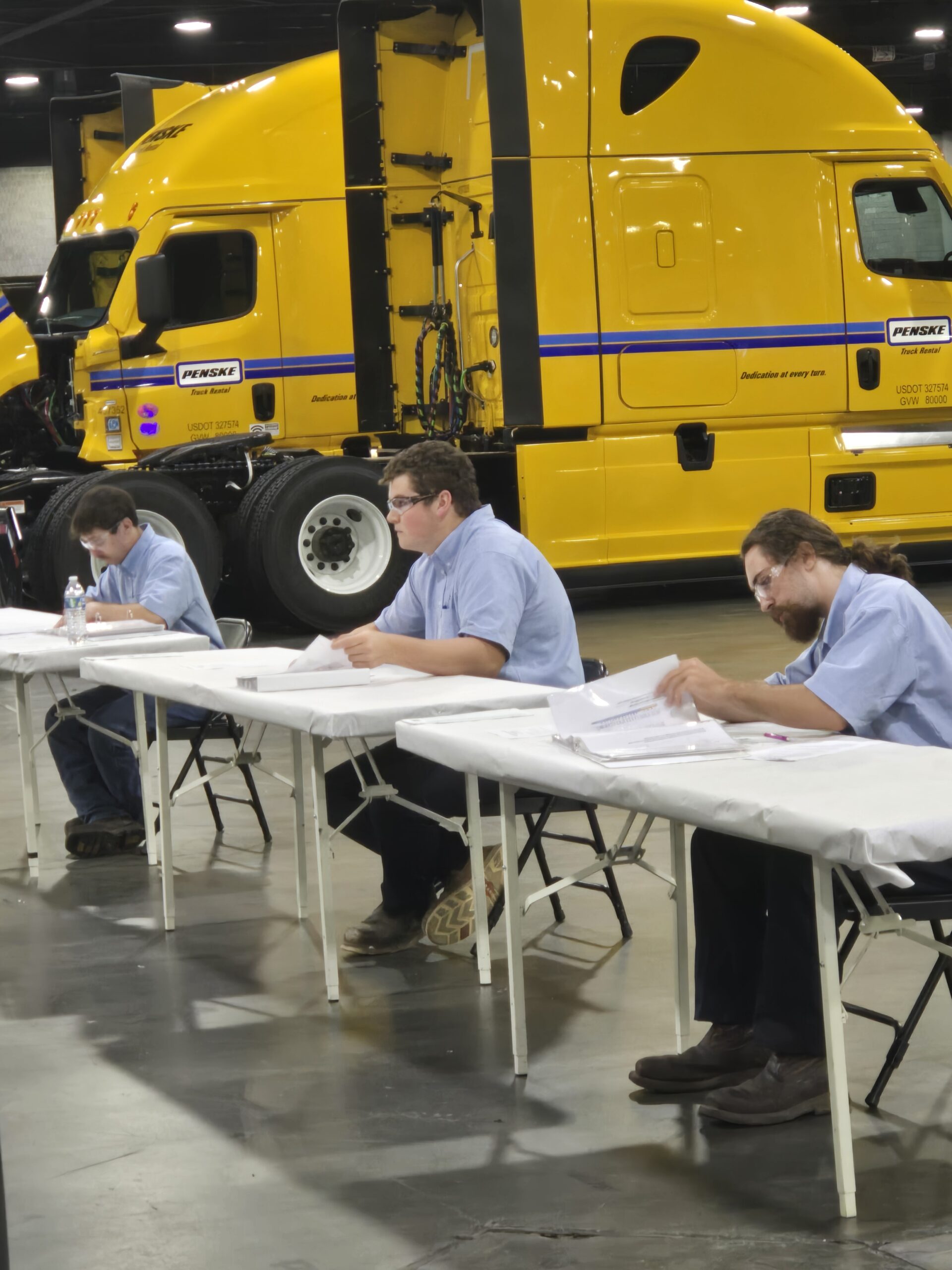 3 men sit at a table, each wearing a blue, short-sleeved collared shirt and dark pants. Behind them is a very large, yellow piece of heavy equipment.